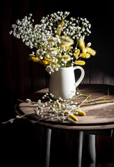 White jug with flowers in the dark photo style, on a wooden table.