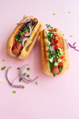 Delicious freshly cooked hot dogs with sausages, vegetables, cheese and sauce on the pastel pink backgound.
