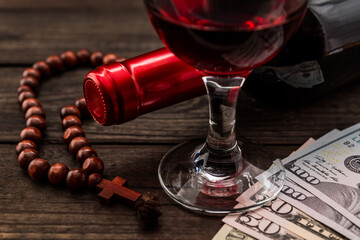 Bottle of red wine with a glass of red wine and money with rosary on an old wooden table. Close up view, focus on the glass of red wine