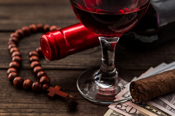 Bottle of red wine with a glass of red wine and money with rosary and cuban cigar on an old wooden table. Close up view, focus on the glass of red wine