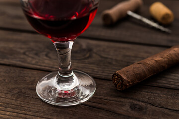 Glass of red wine and cuban cigar with corkscrew and corks on an old wooden table. Close up view, focus on the cuban cigar