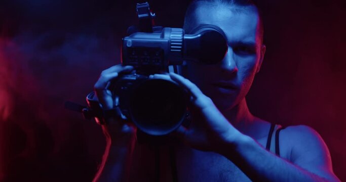 A stylish male hipster cameraman from the 80s with a big retro camera in his hands turns around and points the camera at the viewer. Cyberpunk fashion concept