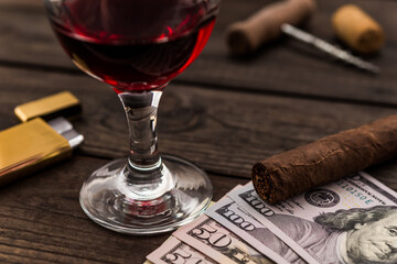 Glass of red wine with money and cuban cigar with corkscrew and corks on an old wooden table. Close up view, focus on the cuban cigar