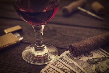 Glass of red wine with money and cuban cigar with corkscrew and corks on an old wooden table. Close up view, focus on the cuban cigar
