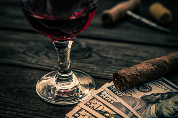 Glass of red wine with money and cuban cigar with corkscrew and corks on an old wooden table. Close up view, focus on the cuban cigar
