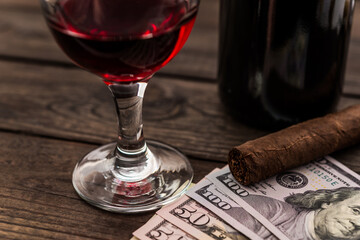Bottle of red wine with a glass of red wine and cuban cigar with lot of money on an old wooden table. Close up view, focus on the cuban cigar