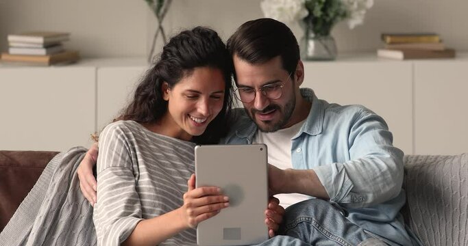 Smiling millennial family couple resting on cozy sofa, using touchscreen pad indoors, web surfing information online, watching funny photo video content in social network, people and modern tech.