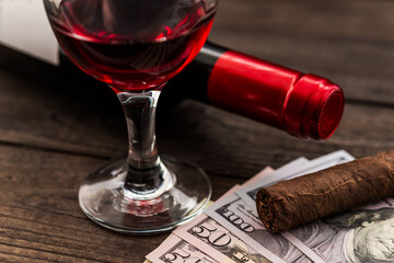 Bottle of red wine with a glass of red wine and cuban cigar with lot of money on an old wooden table. Close up view, focus on the cuban cigar