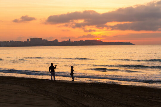 Silhouette of a young couple taking pictures with the sun rising at dawn on the Postiguet Beach in Alicante. Spain