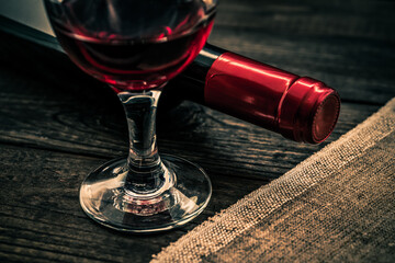Bottle of red wine with a glass of red wine and piece of canvas on an old wooden table. Close up view, focus on the glass of red wine