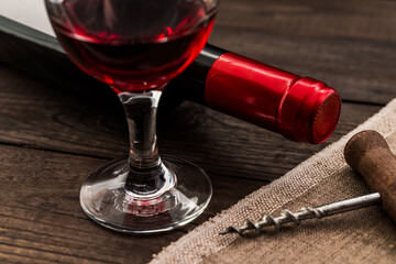 Bottle of red wine with a glass of red wine and piece of canvas with corkscrew on an old wooden table. Close up view, focus on the glass of red wine