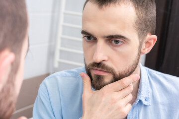 Fototapeta premium Young bearded man looking himself in bathroom mirror, portrait, close-up