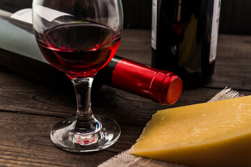 Two bottles of red wine with a glass of red wine and a piece of parmesan on an old wooden table. Angle view, focus on the bottle of red wine