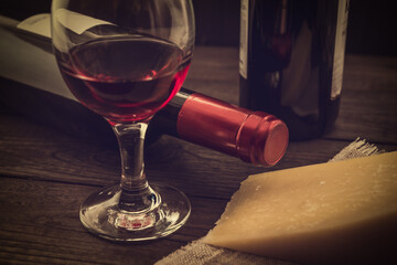 Two bottles of red wine with a glass of red wine and a piece of parmesan on an old wooden table. Angle view, focus on the bottle of red wine