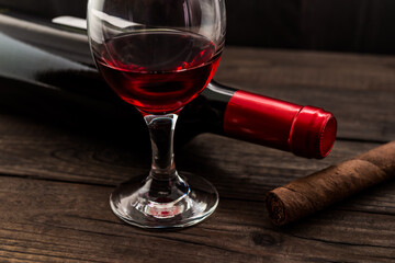 Bottle of red wine with a glass of red wine and cuban cigar on an old wooden table. Angle view, focus on the cuban cigar