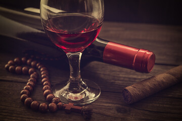 Bottle of red wine with a glass of red wine and cuban cigar with rosary on an old wooden table. Angle view, focus on the cuban cigar