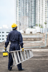 Asian maintenance worker man with protective suit and safety helmet carrying aluminium step ladder and tool box at construction site. Civil engineering, Architecture builder and building service