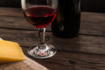 Bottle of red wine with a glass of red wine and a piece of parmesan on an old wooden table. Angle view, focus on the glass of red wine