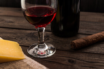 Bottle of red wine with a glass of red wine and a piece of parmesan and cuban cigar on an old wooden table. Angle view, focus on the glass of red wine