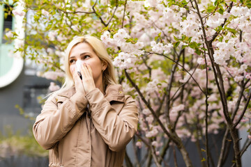 Young pretty woman sneezing in front of blooming tree. Spring allergy concept