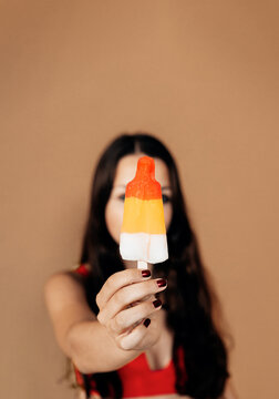 focus on the hand of a brunette girl holding a popsicle. defocused young woman giving the ice pop to the viewer on brown background. vertical picture.