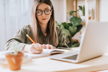Thinking Freelance woman in eye glasses with pencil write in note book, typing at laptop and...