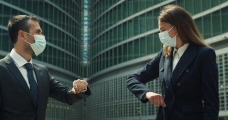 Cinematic shot of business partners or colleagues with protective medical masks greeting by striking with their elbows to respect social distance on skyscrapers background during coronavirus pandemic. - Powered by Adobe