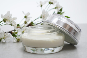 Fototapeta na wymiar .Beauty cream in a glass jar on a light gray background. Decorated with white spring flowers. Unbranded skincare product. Cosmetic cream. Close up, selective focus, side view.