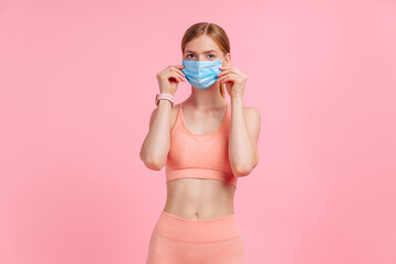 Young fitness woman in medical protective mask, wearing sportswear for workouts, on pink background