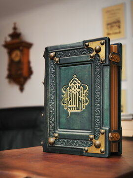 Aged green leather book with gilded calligraphy on the front cover captured in stand up position. 
English translation of the text is: In the name of God, the Most Gracious, the Most Merciful.