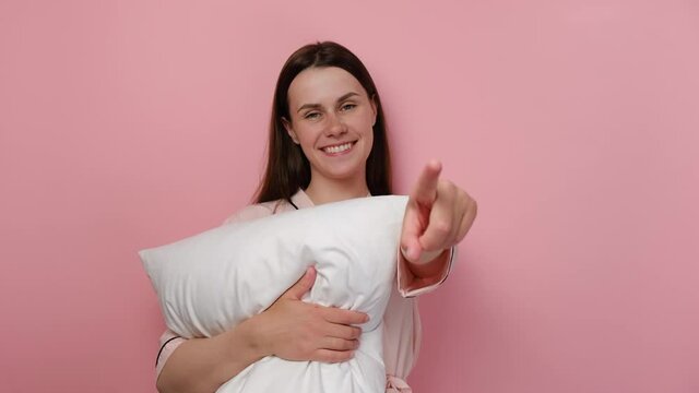 Portrait of beautiful young woman wearing pajama holding white pillow over isolated pink background pointing with finger to camera and to you, hand sign, positive and confident gesture from front
