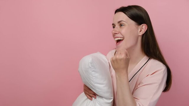 Side view of shocked excited young woman wearing pajama standing over pink isolated background celebrating surprised and amazed for success with arms raised, holding white pillow. Winner concept