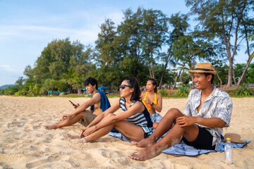 Group of Happy Asian man and woman friends sitting on the beach enjoy picnic and talking together at summer sunset. Male and female friendship relax and having fun summer outdoor lifestyle activity.
