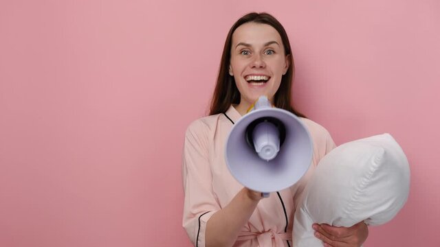 Fun crazy young brunette woman in pajamas shouting through megaphone to announce something, holding white pillow, posing isolated on pastel pink background studio. People emotion lifestyle concept