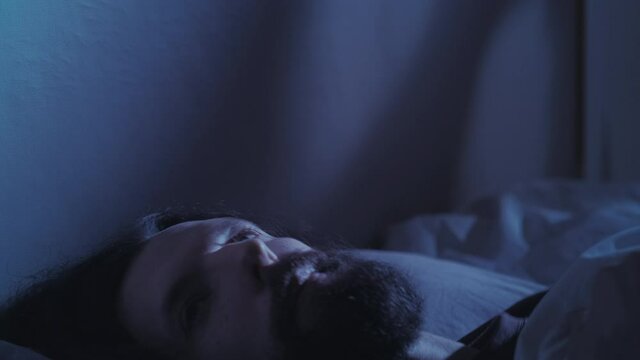 Night insomnia. Trouble sleeping. Anxiety fatigue. Tired exhausted pensive worried man overthinking lying awake alone in bed in dark bedroom with blue moonlight.