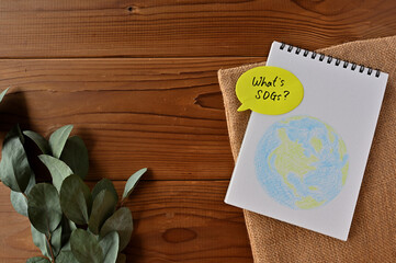 There are eucalyptus leaves and a burlap bag on a wooden table. On top of it is a sketchbook with an illustration of the earth and a speech bubble-shaped sticky note that has 