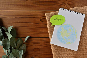 There are eucalyptus leaves and a burlap bag on a wooden table. On top of it is a sketchbook with an illustration of the earth and a speech bubble-shaped sticky note that has 