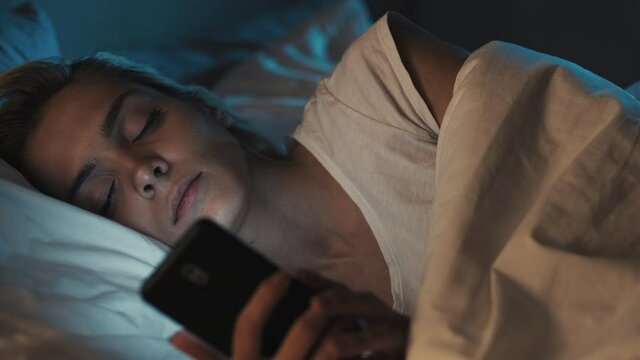 Night online. Bedtime habit. Mobile leisure. Insomnia fatigue. Tired exhausted woman falling asleep with phone dropping device down lying in cozy bed in dark bedroom.