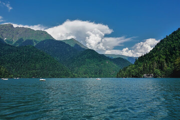 The alpine lake Ritsa is surrounded by mountains. On the slopes there is a coniferous forest. White boats float in the turquoise water. There are picturesque clouds in the azure sky. Summer day.