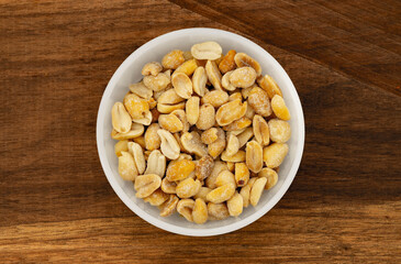 Salted peanuts in white bowl, photographed overhead on dark wood with natural light. Top view of bowl with salty peanut.