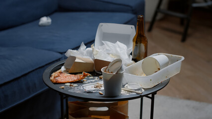 Close up of leftover food on table in empty messy living room, bottle of beer and napkins on blue...