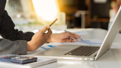 A business woman sitting at a desk and using a smartphone On the desk there is a laptop computer and in the office charts and graphs.