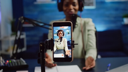 Close up of smartphone recording black woman influencer talking looking into it during online podcast. Social media content creator streaming online broadcast, blogger discussing wearing headphones.