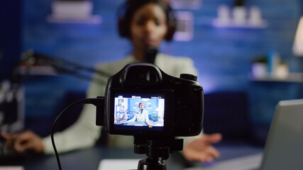 African influencer recording blog looking at professional camera at online talk show home studio. Social media content creator streaming online broadcast, blogger discussing wearing headphones.