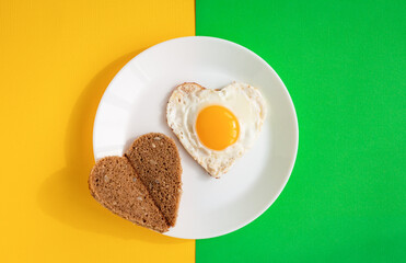 top view of heart-shaped scrambled eggs and toasts, symbol of love, single fried egg on white plate on green and yellow background, romantic minimalistic template, healthy breakfast for sweetheart