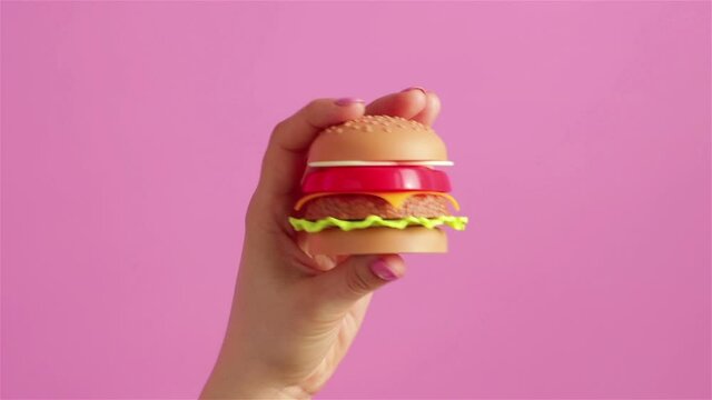 woman hand holding a plastic tiy burger on pink background close up