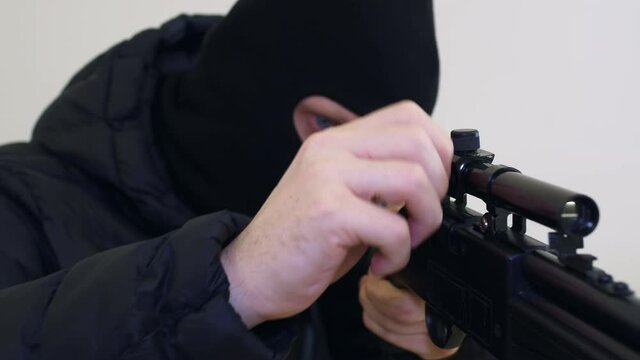 A killer in a black balaclava adjusts a sniper rifle, makes adjustments, twitches the bolt of the rifle and takes aim at the victim. Close-up of weapons.