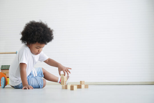 Little Cute African American Boy Sitting On The Floor And Playing Toys Holding Wooden Blocks At Home On White Background