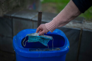 hand throws dirty mask into blue trash can, pollution of city parks with used masks, concept of cleaning the city from plastic masks after quarantine, fight for the environment