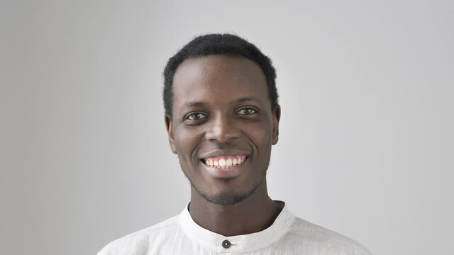 Attractive young African-American man in stylish white shirt smiles looking in camera on light grey background close view